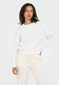 Felpa bianca corta con collo rotondo e spalle scese, abbinata a pantaloni della tuta beige con cordino. Texture morbida e liscia.