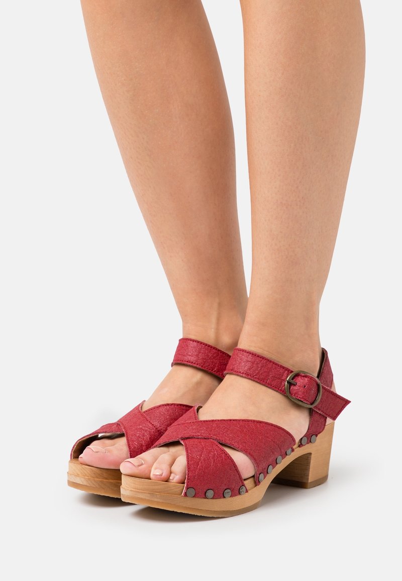 Pieds chaussés de sandales rouges à bout ouvert avec talons en bois, brides à la cheville et clous métalliques le long des bords de la semelle sur un fond blanc.