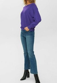 Lila Pullover mit langen Ärmeln und lockerem Schnitt, kombiniert mit blauen ausgestellten Jeans und schwarzen Stiefeletten. Glatte Textur und lässiges Design.