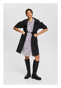 Abrigo negro impermeable con capucha, cinturilla ajustada y bolsillos laterales encima de un vestido floral en tono púrpura claro. La modelo lleva botas negras hasta la rodilla.