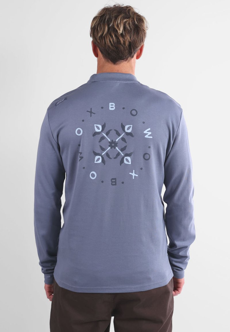 Chemise à manches longues en coton bleu doux avec un design graphique circulaire au dos, présentant du texte et des motifs de feuilles dans des couleurs sombres et claires.