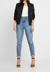 Giacca bomber nera, top bianco a coste, jeans blu sbiaditi a vita alta e décolleté neri con cinturini. Outfit casual con un fit rilassato.