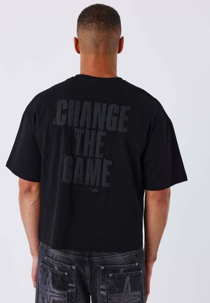 Uomo che indossa una t-shirt nera con la scritta "CHANGE THE GAME" sulla schiena, abbinata a jeans grigio scuro con tasche cucite.