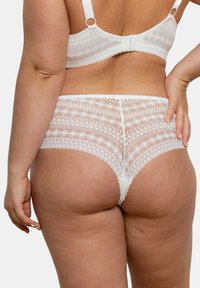 String blanc en dentelle avec des motifs géométriques et des bords festonnés, doté d'une texture douce et d'une coupe mi-haute.