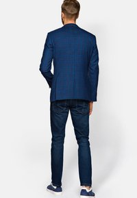 Męska niebieska marynarka w kratę o dopasowanym kroju, z rozcięciem z tyłu i kontrastowym wzorem; zestawiona z ciemnymi dżinsami i niebieskimi trampkami.