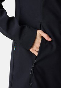 Czarna kurtka softshell z ręką w bocznej kieszeni, widocznymi szwami oraz małą naszywką ze szwedzką flagą w pobliżu szwu.
