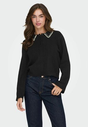 Vrouw in een zwart kabelgebreid sweater met een parelversierde kraag en donkerblauwe spijkerbroek, staand met één hand in de zak tegen een witte achtergrond.