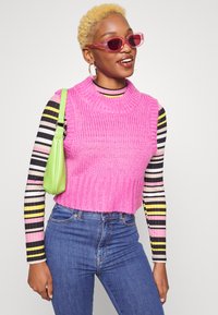 Sweater sem mangas rosa de malha com bainha canelada, sobre uma camisa de manga longa às riscas. Os acessórios incluem uma bolsa verde e óculos de sol rosa.