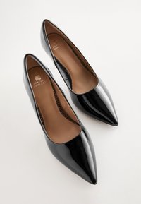 Zwarte hoge hakken van lakleer met puntige neuzen en een gladde afwerking. De binnenkant heeft een tan lederen voetbed met ingebrand merk.