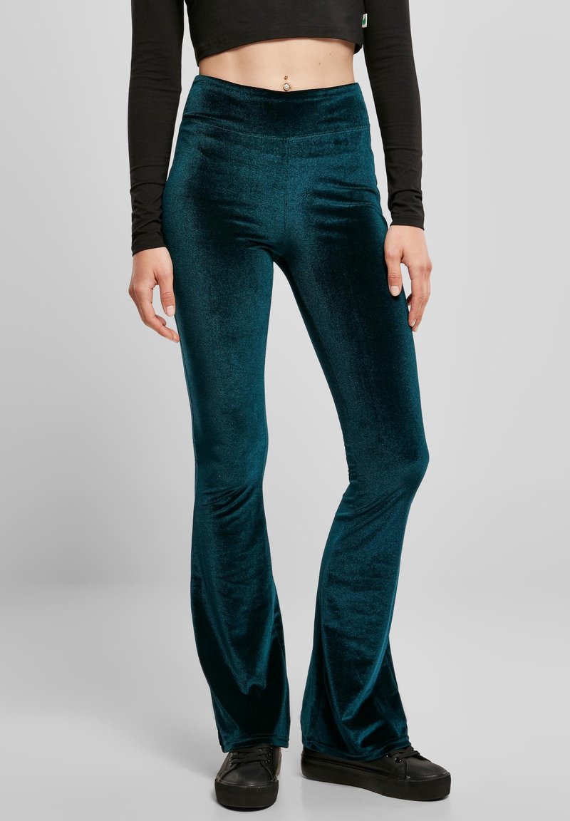 Femme portant un pantalon évasé en velours bleu sarcelle, un crop top noir à manches longues, des baskets noires et un piercing au nombril, debout devant un fond gris.