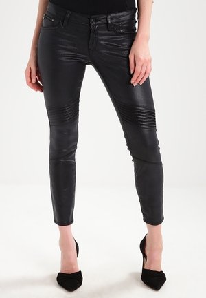 Woman wearing black textured leather pants with horizontal ribbed detailing on thighs, paired with black high-heeled shoes, standing against a white background.