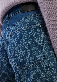 Κοντινή λήψη μπλε τζιν με υφή και στίγματα και πίσω τσέπη, φορεμένα με μωβ πλεκτό πουλόβερ.