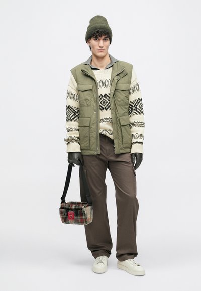 Olive zelen puffer brezrokavnik čez krem pulover z črnimi geometrijskimi vzorci, v kombinaciji s rjavimi hlačami in belimi teniskami; nosi karo torbo.