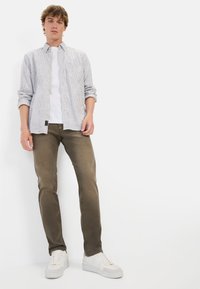 Lichtblauw gestreept overhemd met knopen; witte T-shirt eronder; olijfgroene broek; witte sneakers met een gestructureerde zool. Eenvoudige, casual outfit.