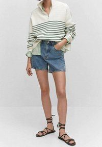 Weißes, geripptes Langarmoberteil mit grünen Streifen, kombiniert mit blauen Jean-Shorts und schwarzen Riemchensandalen. Lässig, entspannte Passform.