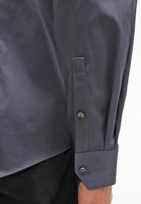 Maglietta grigia a maniche lunghe con una texture liscia, dotata di un polsino con bottone e discreti dettagli di cucitura sulla manica e sul colletto.