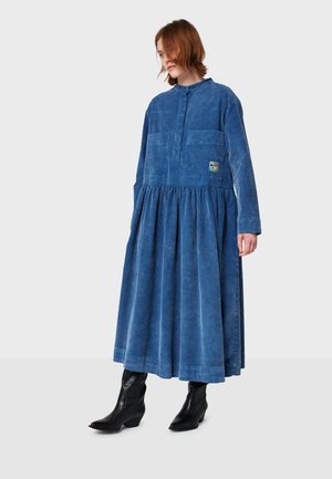 Blaue Jeanskleid mit langen Ärmeln, durchgehender Knopfleiste, zwei Brusttaschen, gerafftem Rock und einem bunten Patch auf der linken Seite. Getragen mit schwarzen Stiefeln.