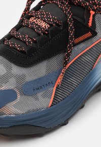 Scarpa sportiva con tomaia in mesh grigia e nera, dettagli arancioni e suola in gomma nera con motivo. Presenta l'elemento di design "PWR TAPE".