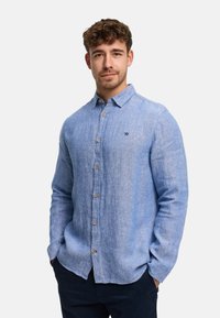 Nuori mies, jolla on kiharat hiukset, pukeutuneena vaaleansiniseen, ruskeilla nappeilla varustettuun nappipaitaan ja tummiin housuihin, kädet taskuissa, neutraali tausta.