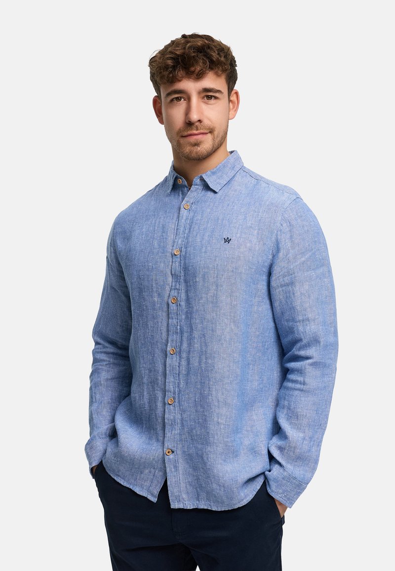 Nuori mies, jolla on kiharat hiukset, pukeutuneena vaaleansiniseen, ruskeilla nappeilla varustettuun nappipaitaan ja tummiin housuihin, kädet taskuissa, neutraali tausta.