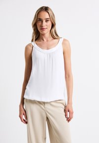 Top sin mangas blanco con un escote redondeado y detalles recogidos. Hecho de una tela suave. Combinado con pantalones de pierna ancha de color beige claro.