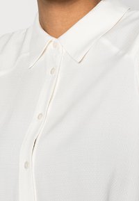 Camicia bianca con colletto, realizzata in un tessuto testurizzato con un motivo sottile, caratterizzata da una chiusura a bottoni e un colletto liscio e impeccabile.