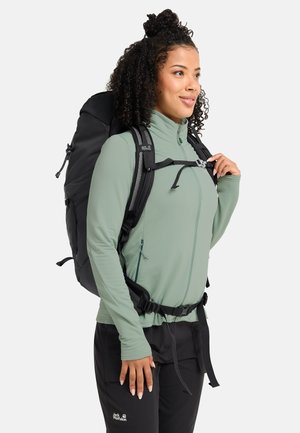 Vrouw die de borstband van een zwarte rugzak verstelt, met een lichtgroene jas en zwarte outdoorbroek, tegen een witte achtergrond.