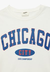 Biała koszulka z okrągłym dekoltem z niebieskim i czerwonym napisem "CHICAGO" oraz "CITY" w odważnej czcionce, a poniżej w mniejszym niebieskim tekście "STATE CHAMPIONSHIP".