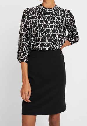 Kobieta ubrana w czarno-białą bluzkę z geometrycznym wzorem na długim rękawie, włożoną do gładkiej czarnej spódnicy o długości do kolan.