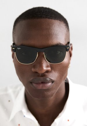 Zwarte Ray-Ban zonnebril met gouden accenten, met een rechthoekige vorm en donkere lenzen, gedragen door een persoon in een wit shirt met kleurrijke spikkels.