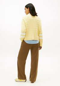 Jersey de lana amarillo con un patrón texturizado, rayas blancas en las mangas, superpuesto a una camisa de rayas azules, combinado con pantalones marrones.