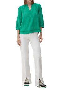 Grün strukturierte Bluse mit Dreiviertel-Ärmeln und V-Ausschnitt, kombiniert mit weißen, ausgestellten Hosen mit seitlichen Schlitzen und schwarzen Sneakers.