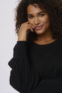 Pull noir avec des manches plissées à côtes et un col rond. Le mannequin touche délicatement son visage, mettant en valeur le design du vêtement.