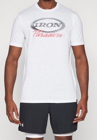 Biała bawełniana koszulka z krótkim rękawem, z czarnym owalnym logo z napisem "IRON" i "Paradise" w różowym kolorze, zestawiona z ciemnymi szortami.