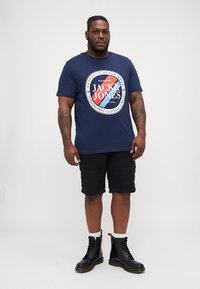 Maglietta blu navy con un logo circolare bianco e rosso sul davanti. Abbinata a pantaloni cargo neri e stivali neri con lacci.