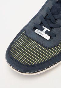 Zapatilla azul marino con parte superior de malla que presenta acentos amarillos, etiqueta de logo plateada y costuras blancas en contraste sobre una suela flexible.