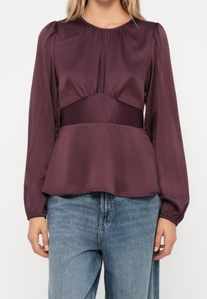 Femme portant un chemisier en satin violet à manches longues avec des épaules bouffantes et une large ceinture, associé à un jean bleu.