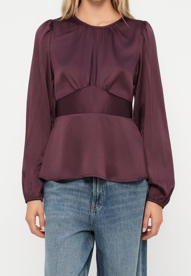 Femme portant un chemisier en satin violet à manches longues avec des épaules bouffantes et une large ceinture, associé à un jean bleu.