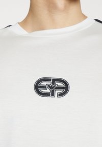 Vit kortärmad skjorta med rund halsringning, med en svart broderad logotyp på bröstet och svarta detaljer på axlarna.