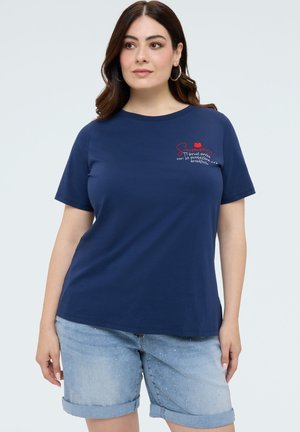 Mujer que lleva una camiseta azul marino con letras rojas y pantalones cortos de mezclilla azul claro con puños doblados y pequeños adornos.