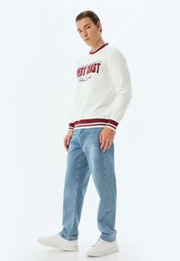 Felpa bianca con testo "West Coast" in bordeaux, polsini e scollatura a costine. Un paio di jeans azzurri a gamba dritta e sneakers bianche completano il look.