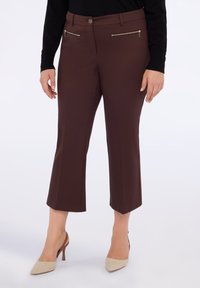 Femeie îmbrăcată în pantalonii scurți maro cu buzunare frontale cu fermoar și tocuri bej, stând în fața unui fundal alb.