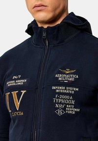 Giacca in pile blu navy con zip, caratterizzata da testo ricamato in oro e bianco, inclusi loghi militari e un design di volo.