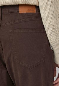 Braune Hose mit einer aufgenähten Tasche auf der Rückseite, und ein beiger Strickpullover, dessen Ärmel und Hand seitlich sichtbar sind.