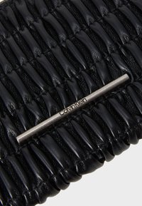 Pochette en cuir noir avec texture striée, dotée d'une barre d'accent métallique argentée portant le logo "Calvin Klein".
