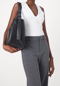 Černá kožená kabela ve tvaru kbel�íku s plochým dnem a nastavitelným popruhem, držena vedle šedých strukturovaných upravených kalhot a bílého topu s výstřihem do V.