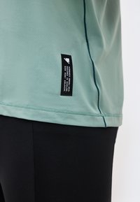 Φόρεμα από ανοιχτό πράσινο υφασμάτινο πουκάμισο με μαύρη ορθογώνια ετικέτα στο πλάι, φορεμένο πάνω από μαύρο εφαρμοστό παντελόνι.