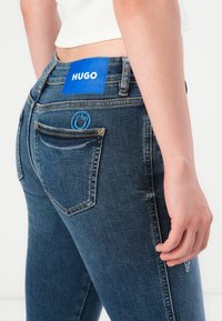 Detaljni prikaz osobe koja nosi tamnoplave Hugo traperice s plavom Hugo oznakom i detaljem nasmijanog lica na stražnjem džepu, uz vidljivu bijelu majicu.