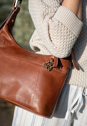 Brown leather shoulder bag with a smooth texture, featuring a decorative metal accent and a wide strap, positioned against a light sweater.