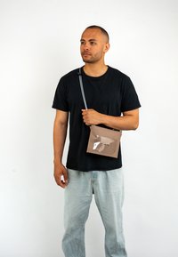 Borsa a tracolla beige con tasca frontale trasparente, forma rettangolare, materiale in tessuto e tracolla regolabile, tenuta da una persona.